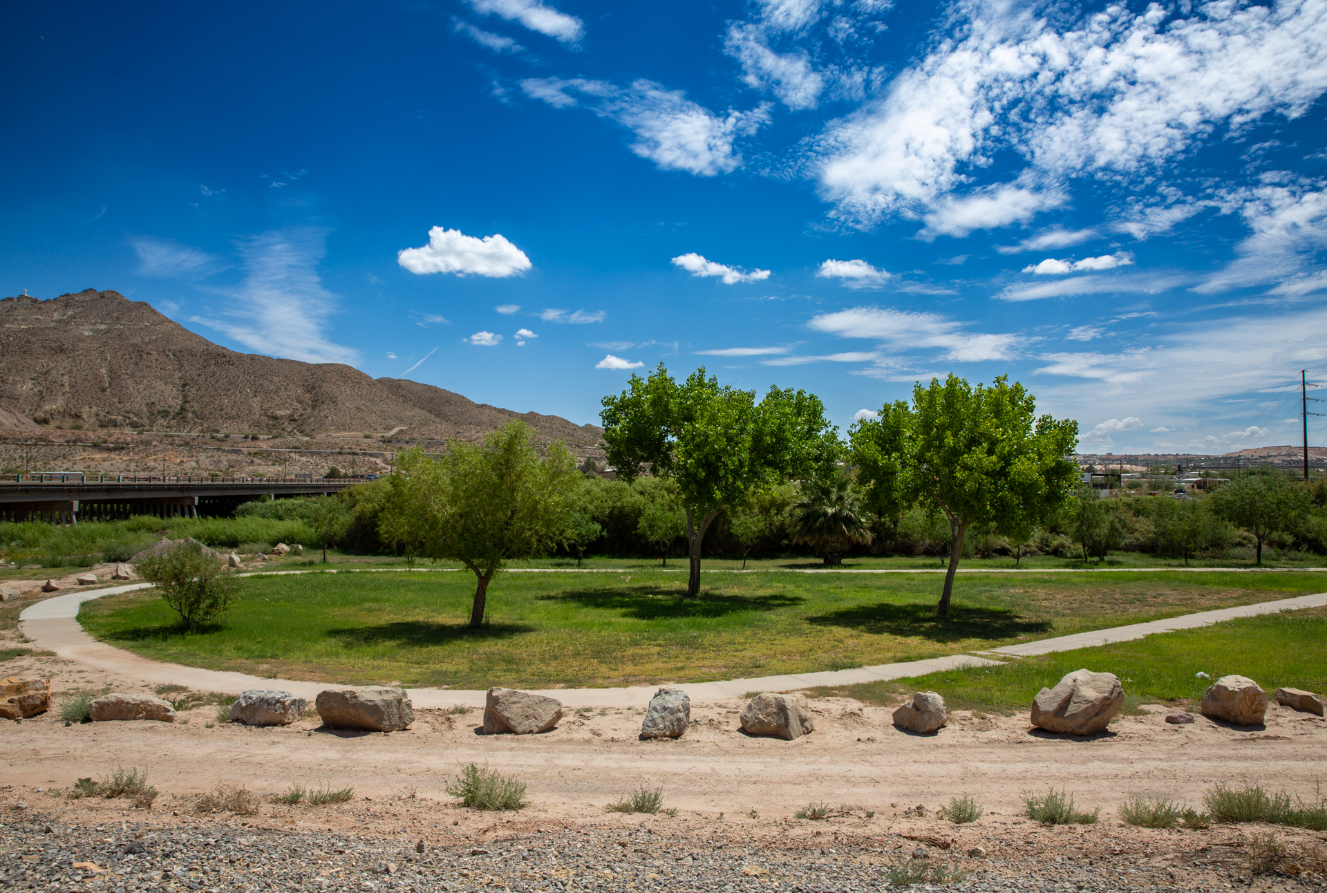 The River Levee Park walking path, grass, and trees under a blue sky with a mountain in the background.