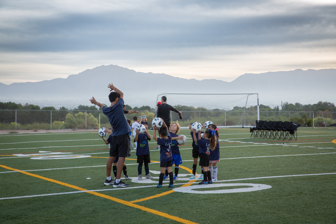 A coach leads young players in a drill, each holding a soccer ball overhead, with the mountains behind them / Un entrenador dirige un ejercicio con los niños, cada uno con su balón en alto y las montañas de fondo